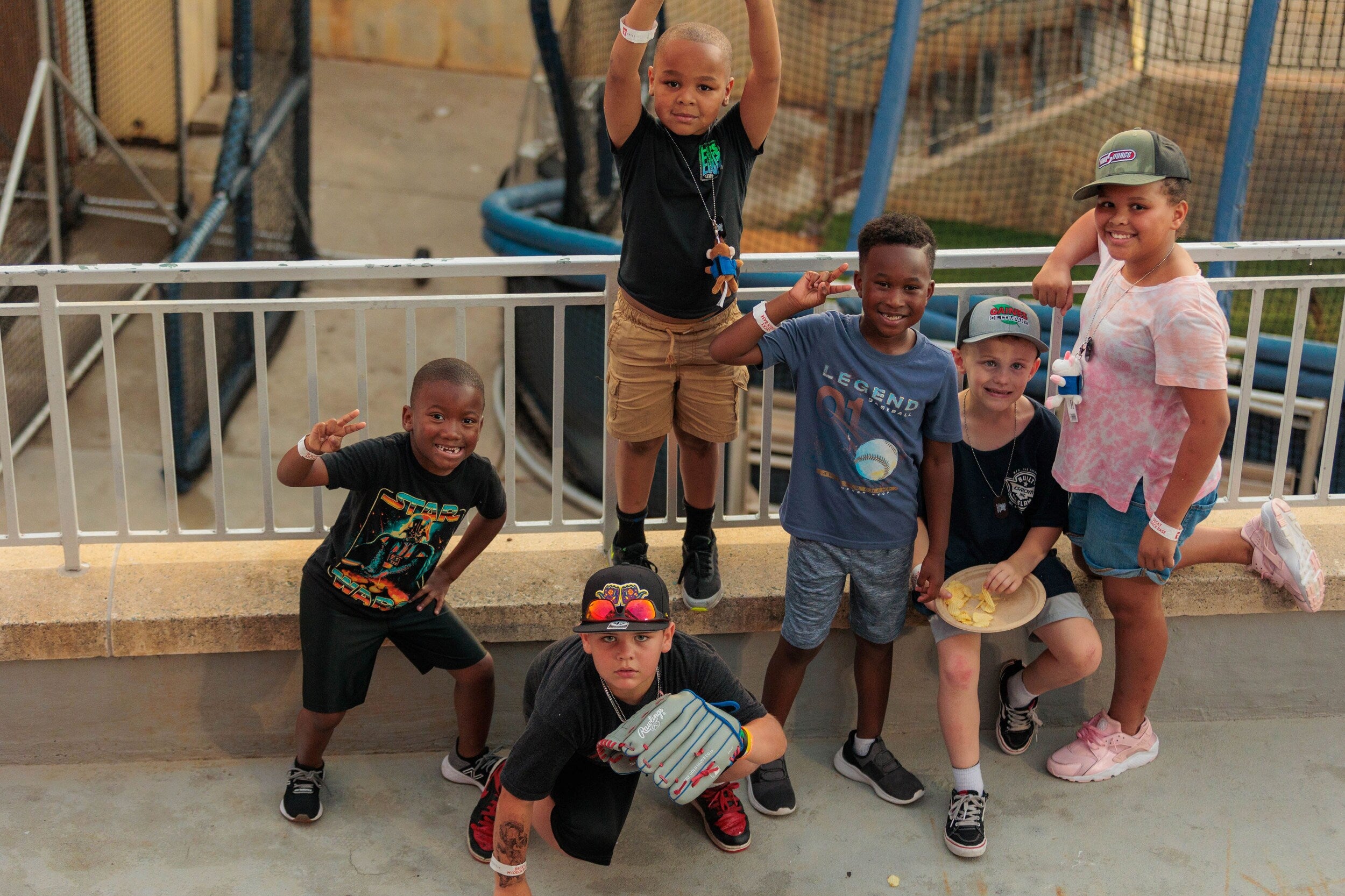 Kids posing together near a stadium railing, showing playful gestures and holding snacks and a baseball glove.