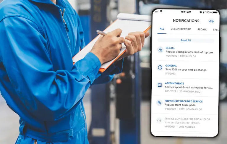 Mechanic writing on a clipboard beside a phone screen showing service and recall notifications
