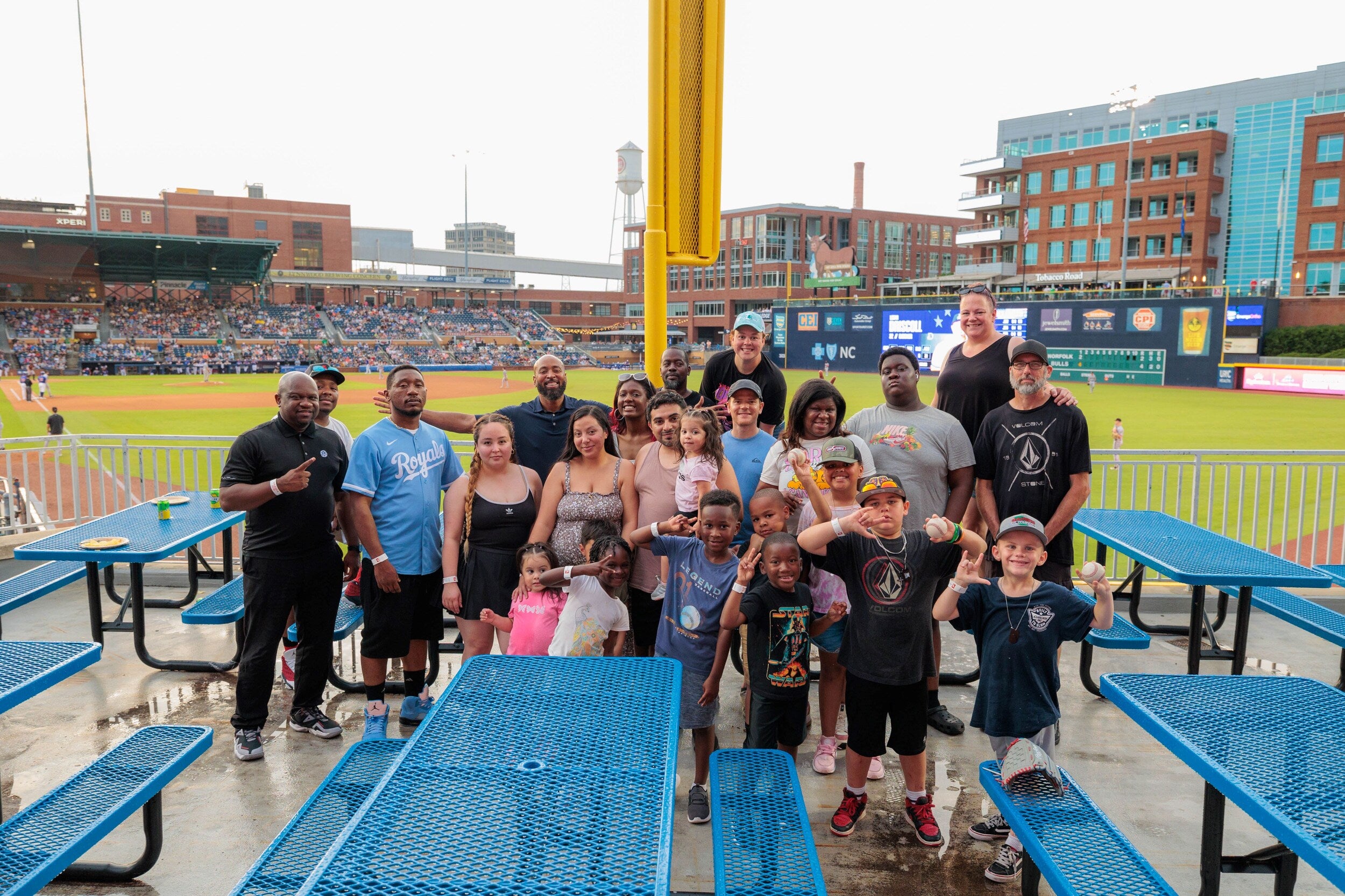 A large group of adults and children posing together at a baseball stadium with the field and stands in the background.