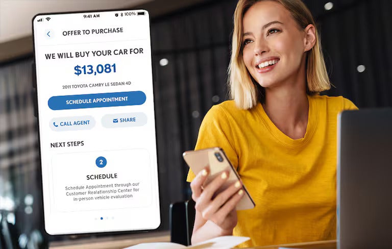 Person holding a smartphone while viewing a car purchase offer screen displayed beside them
