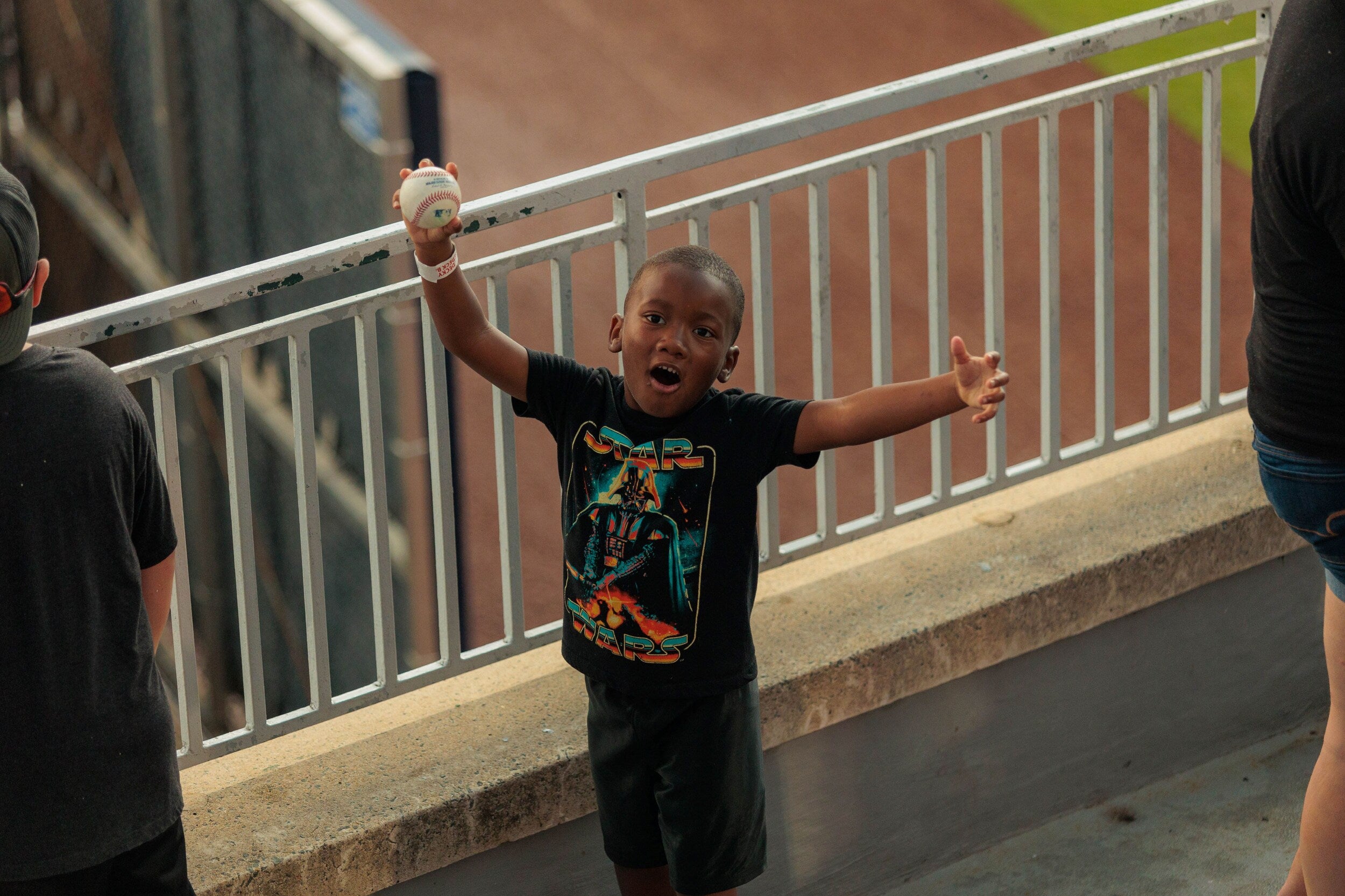 A child standing near a stadium railing holding up a baseball with arms outstretched
